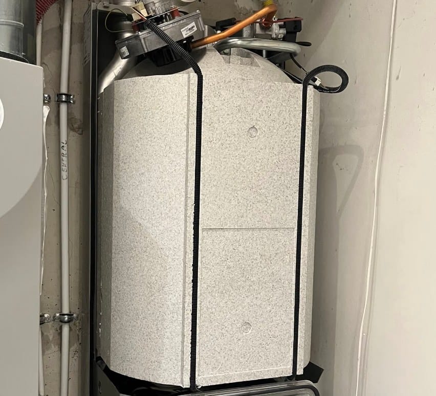 Cv-ketel in een opstelruimte met de mantel nog in transportfolie en spanbanden eromheen. Bovenop zijn de ventilator, het gasblok en de koperen gasaansluiting zichtbaar, met de rookgasafvoer links en de condensafvoer rechts.